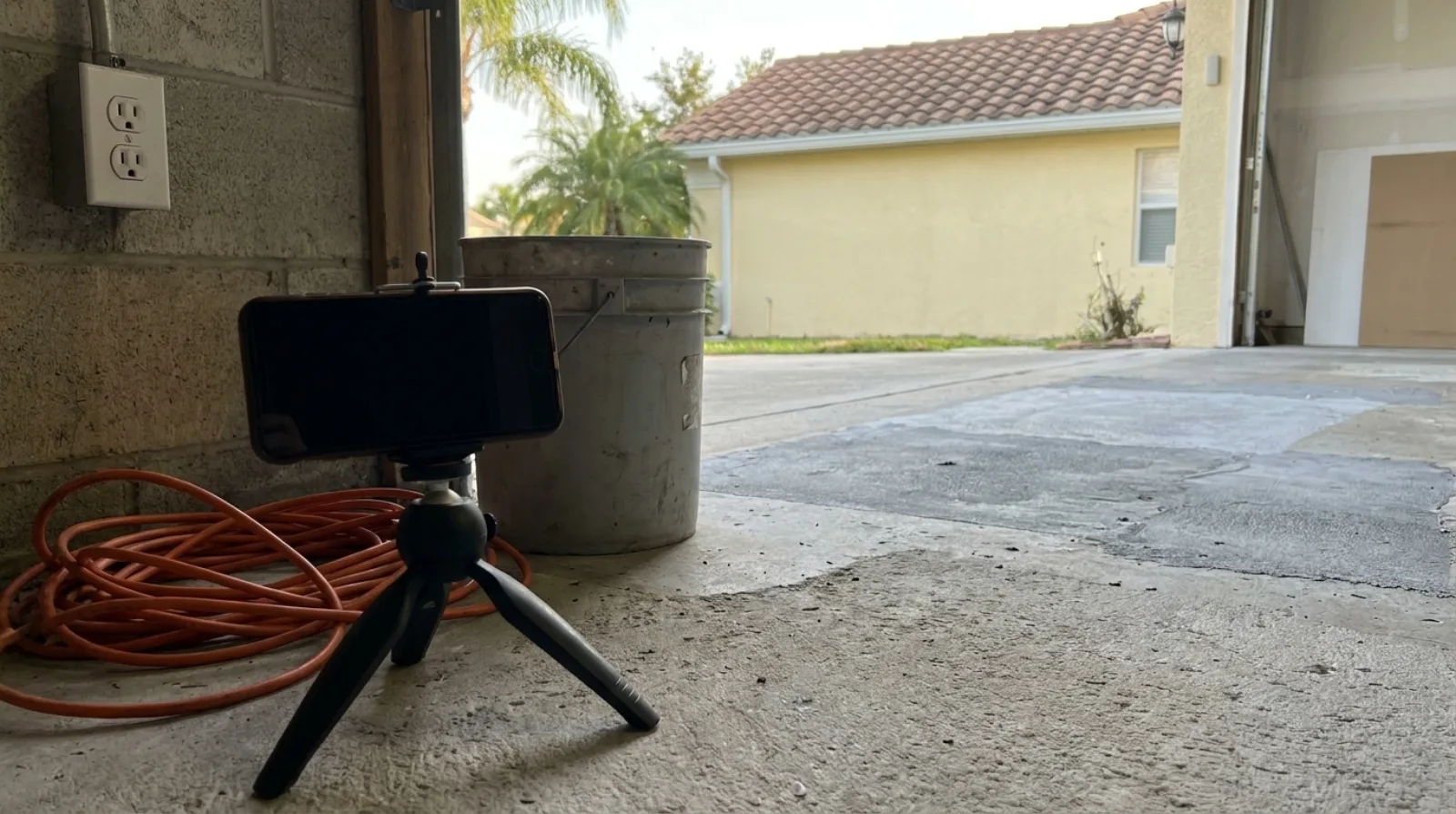 A smartphone clamped to a small black tabletop tripod on the floor of an open Florida garage, aimed at a patched and partially ground concrete slab, with a coiled orange extension cord and a plain plastic pail beside it and a barrel tile roof, palms and pastel stucco house visible through the open door